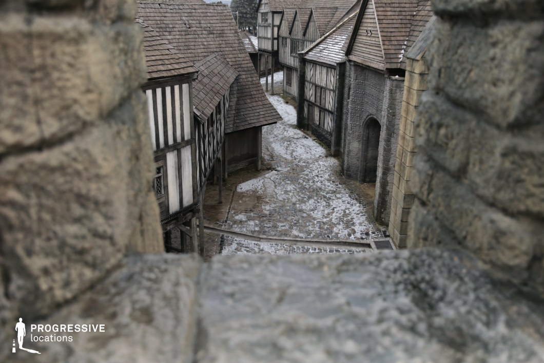 This image captures a winding cobblestone street flanked by half-timbered buildings and stone structures, viewed through a narrow opening in a fortress wall. The tightly packed architecture, steep wooden rooftops, and irregular alleyway evoke the atmosphere of a fortified medieval town. The layered textures of timber and stone, along with the closed-in perspective, create a sense of tension and immersion. This setting is ideal for historical dramas, fantasy epics, or adventure films, serving well for chase scenes, town confrontations, or establishing shots of a period village.