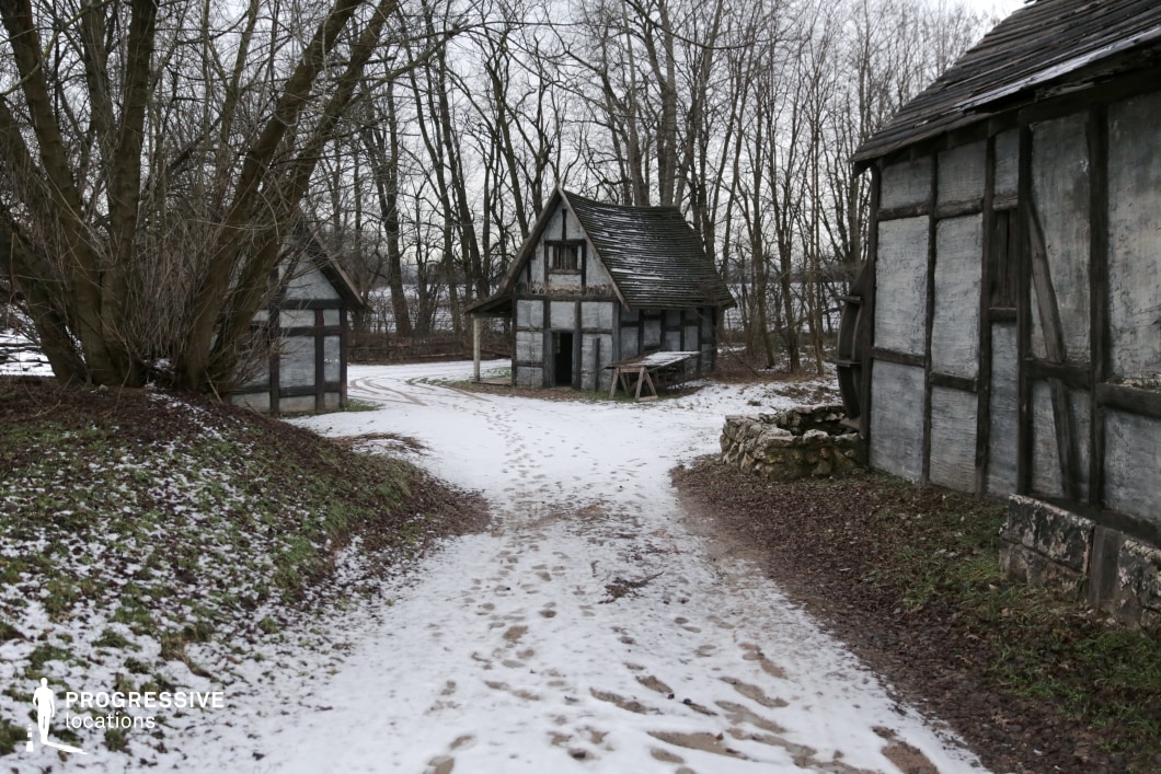 This remote cluster of half-timbered cottages sits along a forked dirt path surrounded by bare trees and gentle embankments, suggesting a modest medieval village or the outskirts of a rural settlement. The simple architecture and sparse layout give the area an authentic, lived-in character, ideal for conveying humble livelihoods or secluded lifestyles. This location is perfectly suited for fantasy, historical drama, or folklore-inspired productions, especially scenes involving village life, secret hideouts, or characters retreating into nature.