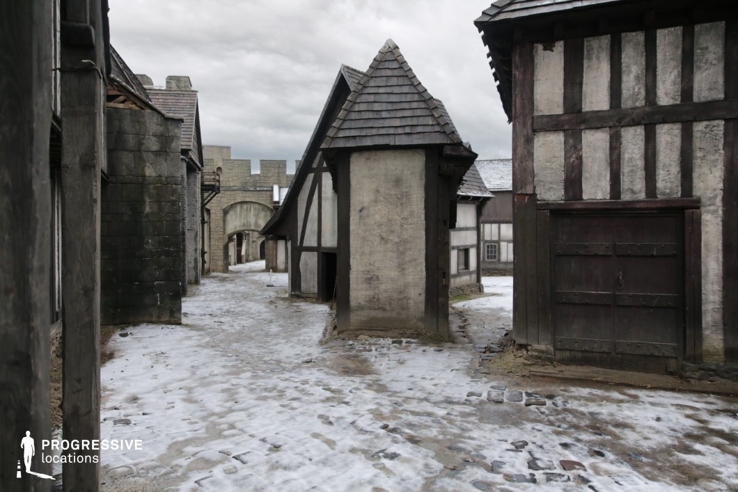 A tight, curving alleyway lined with timber-framed facades and aged stonework creates an immersive medieval village setting. The irregular angles and close spacing of the buildings evoke a sense of organic town growth, while the stone gate in the background anchors the scene with a fortified element. The layout allows for dynamic camera movement and layered depth. This location is especially well-suited for filming fantasy tales, period dramas, or historical thrillers, particularly scenes involving townsfolk life, secretive meetings, or chases through old village streets.