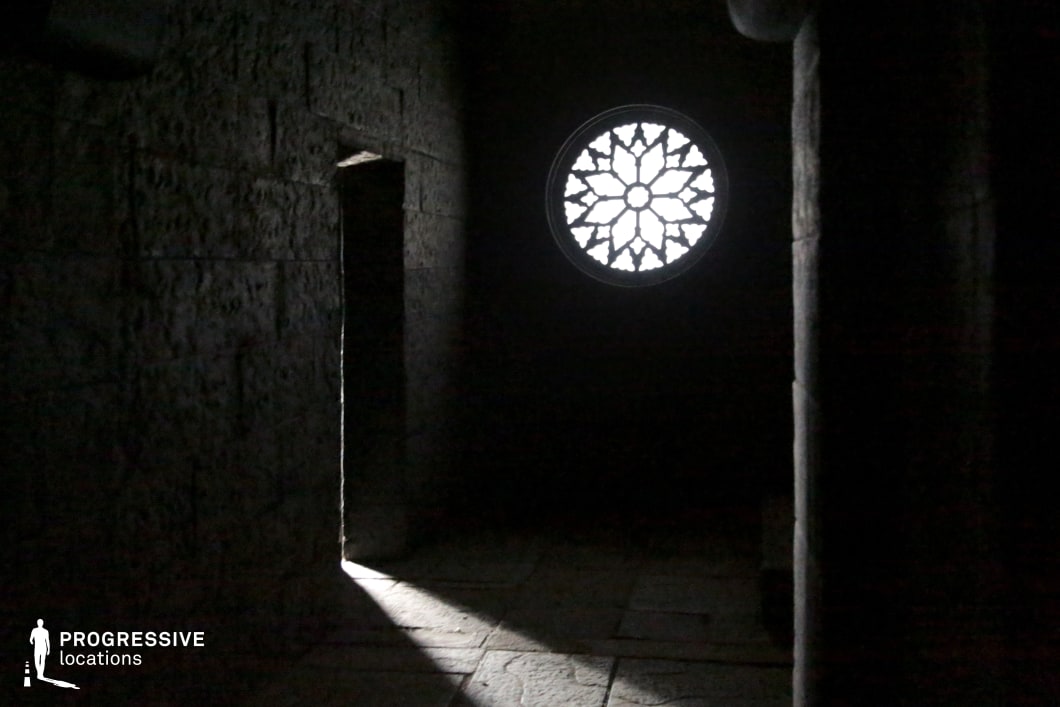 This dimly lit stone corridor features textured masonry and a striking circular rose window that casts a patterned light onto the floor, creating a dramatic and atmospheric interplay of shadow and form. The narrow doorway and austere architecture evoke a sense of secrecy, solemnity, or even confinement, suggesting a historical, religious, or monastic setting. This location is ideal for filming scenes with a mysterious, gothic, or spiritual tone, such as period dramas, fantasy quests, horror sequences, or introspective character moments involving solitude or revelation.