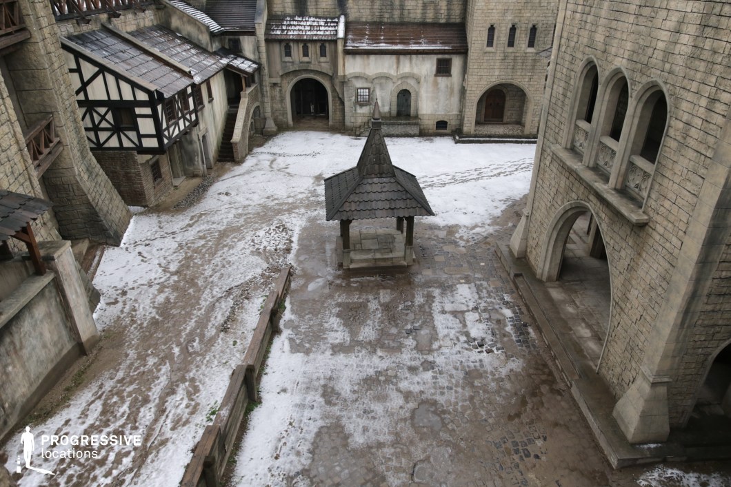 This enclosed courtyard is surrounded by high stone walls, arched passageways, and half-timbered facades, anchored by a prominent central well structure beneath a steep, gabled roof. The worn cobblestones and weathered architecture convey a strong sense of age and function, suggesting a fortified settlement or medieval town square. The layout offers depth, layered elevation, and varied angles ideal for dynamic camerawork. This location is particularly suited for filming period battles, town gatherings, royal proclamations, or fantasy scenes involving court intrigue or public rituals.