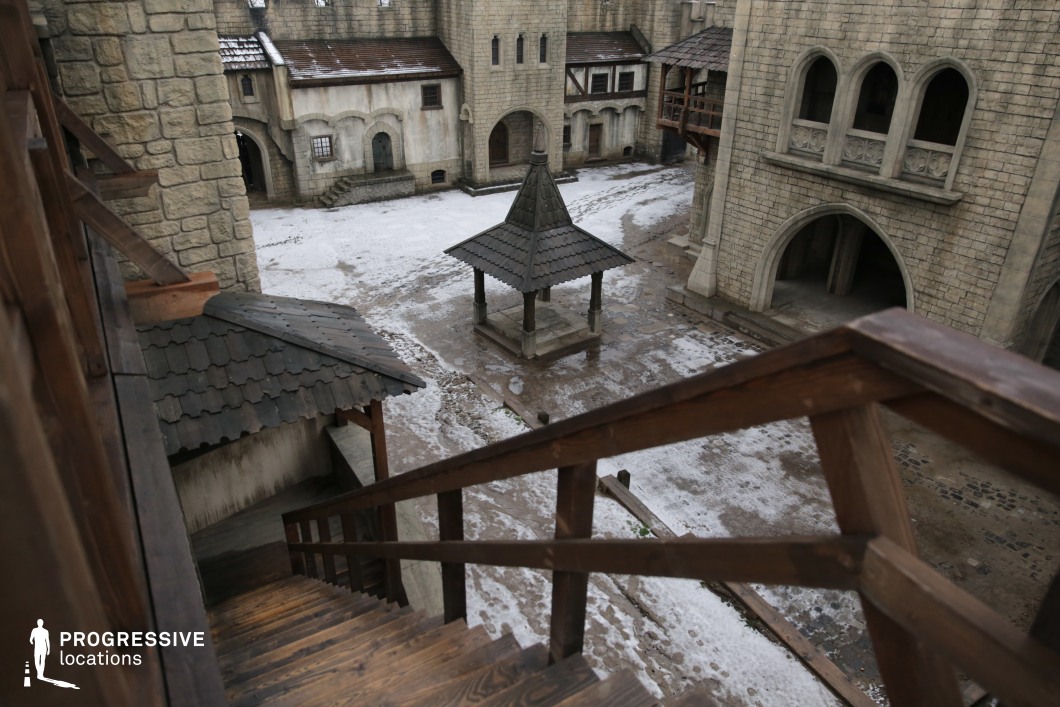 This expansive stone courtyard is framed by fortress-like architecture, including arched entrances, thick walls, and tall buildings with medieval design elements. The central pavilion, roofed with shingles and supported by heavy pillars, adds a focal point and enhances the authenticity of the scene. The wide layout, combined with the vantage point from the wooden stairs, allows for dynamic camera movement and staging. This location is ideal for medieval or fantasy films, particularly for marketplace scenes, public gatherings, confrontations, or dramatic announcements in a fortified town or castle setting.