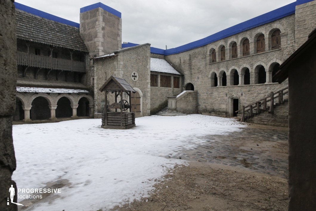 This enclosed stone courtyard features a central wooden well, surrounded by multi-level medieval-style architecture with arched colonnades, narrow windows, and a stair-accessed upper level. The thick masonry walls and robust towers convey a fortified atmosphere, reinforced by heavy wooden shutters and the uniform use of rough-hewn stone. The layout includes a mix of flat and gently sloped surfaces, with a partially snow-covered cobblestone ground offering contrast in texture and tone. The rectangular open space is visually framed by consistent geometric lines and shadowed arcades, enhancing depth and symmetry. This location is ideal for historical, fantasy, or period film productions, offering an authentic setting for castle interiors, fortress courtyards, or medieval town scenes.
