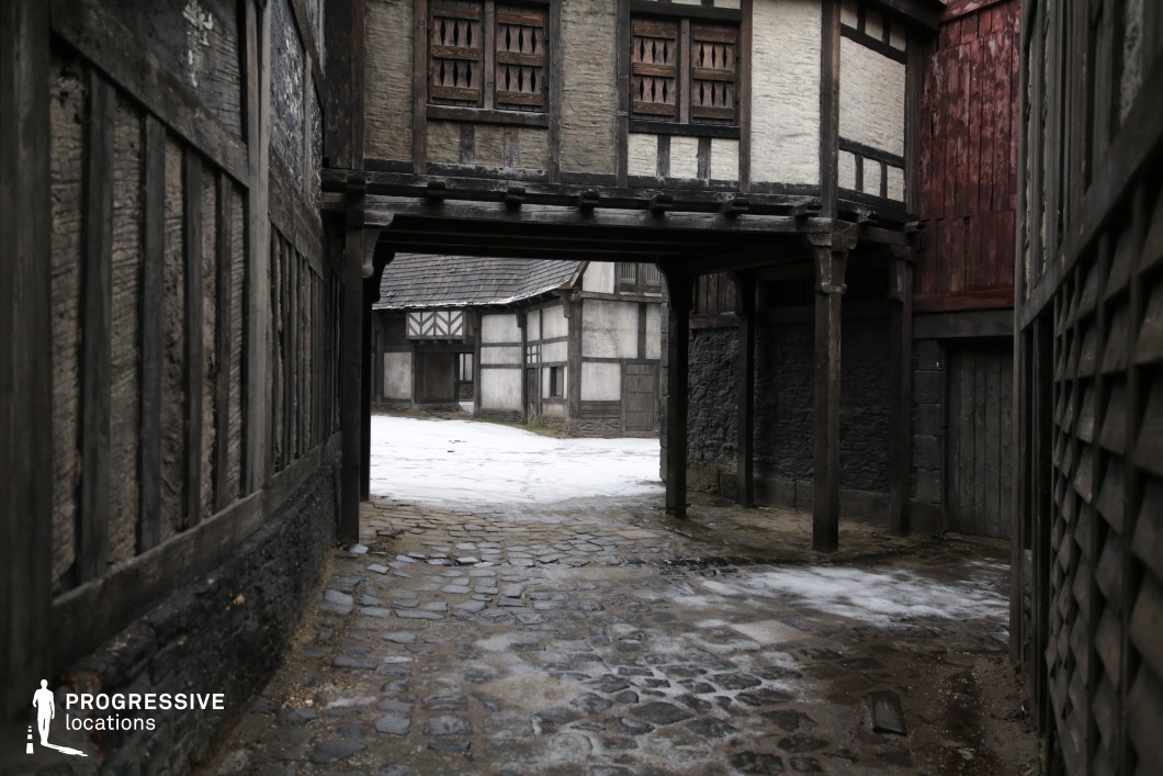 This location features a tight cobblestone alley leading through a low archway into an open square surrounded by half-timbered buildings with weathered wooden facades and irregular architecture. The overhanging upper floors and enclosed space evoke a strong sense of an old European village or fortified town, rich in texture and atmosphere. The dense layout and historical style make it ideal for filming period dramas, fantasy tales, or mystery thrillers, especially scenes involving secret meetings, pursuits, or everyday life in a medieval or early modern setting.
