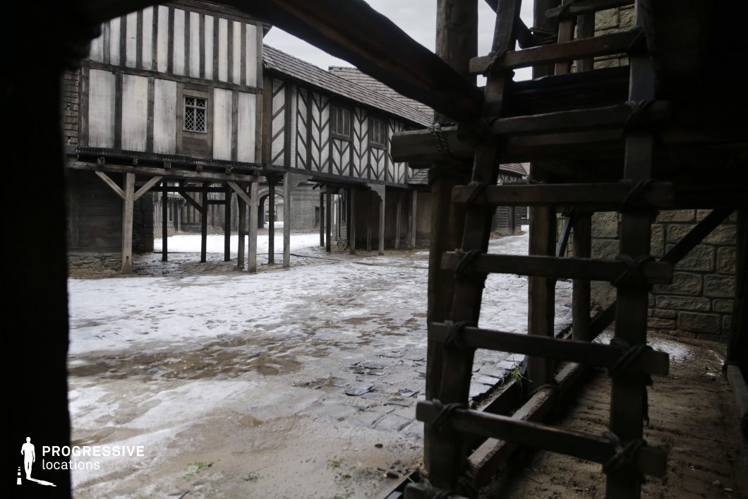 This setting captures a narrow medieval street lined with elevated half-timbered buildings supported by wooden stilts, creating an enclosed and immersive environment. The structures feature rich geometric patterns and period-style detailing, while the foreground includes a rustic wooden staircase and stone walls, adding layers of visual depth. The composition evokes a gritty, lived-in atmosphere, making it ideal for filming darker scenes in historical thrillers, fantasy adventures, or dramatic period pieces involving clandestine meetings, escapes, or urban conflict.
