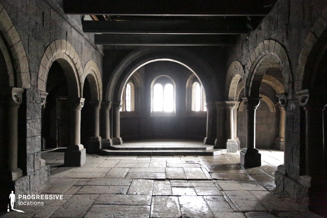 This space features a series of rounded stone arches supported by stout columns, characteristic of Romanesque architecture. The rough-hewn stone floor and heavy masonry walls evoke a sense of antiquity and solemnity, while the deeply recessed windows allow only minimal natural light to filter in, enhancing the cavernous and cloistered atmosphere. The symmetry and repetitive arch forms create a rhythm that feels both monastic and ceremonial. This location is ideal for historical dramas, medieval fantasy, religious or gothic horror genres, serving well as a crypt, dungeon, monastery, or ancient ceremonial chamber.