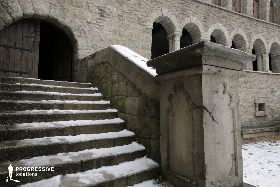 This close-up view captures a weathered stone staircase leading to a large arched doorway, framed by a sturdy, carved parapet and backed by a façade of uniform ashlar masonry with Romanesque-style arcades. The thick stone blocks and robust architectural lines highlight the durability and historical depth of the construction, while the ornamental stonework on the balustrade adds a touch of subdued decoration. The composition emphasizes the vertical progression and textural contrast between rough stone surfaces and defined structural elements. Ideal for period films, medieval dramas, or historical thrillers, this location offers authentic architectural detail and a strong sense of place suitable for transitions, confrontations, or suspenseful entrances.