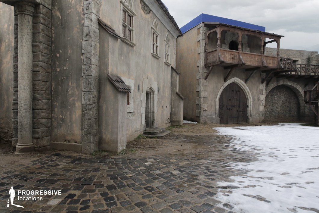 This location features a convincingly weathered medieval-style courtyard, defined by textured stone facades, pointed arched doorways, and aged cobblestone paving. A standout architectural detail is the wooden balcony supported by thick beams, paired with an adjoining stone archway and upper walkway that evokes the atmosphere of an old fortress or town square. The materials and composition suggest a historical European setting, while the simplicity and neutrality of the environment allow for easy adaptation. This location is ideal for filming period dramas, historical fantasy, or medieval adventure scenes—particularly those involving town confrontations, secret meetings, or fortress entrances.