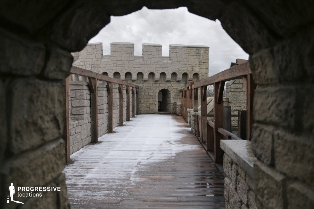 This elevated stone walkway stretches along the inner side of a fortified castle wall, framed by crenellated battlements and wooden railings that enhance its defensive character. The perspective through a stone archway emphasizes depth and enclosure, ideal for tracking shots or tense dialogue scenes. The uniform masonry and minimal adornment contribute to a realistic medieval aesthetic. This setting is ideal for historical war films, fantasy battles, or scenes involving sentries, stealth, or confrontation along the ramparts.