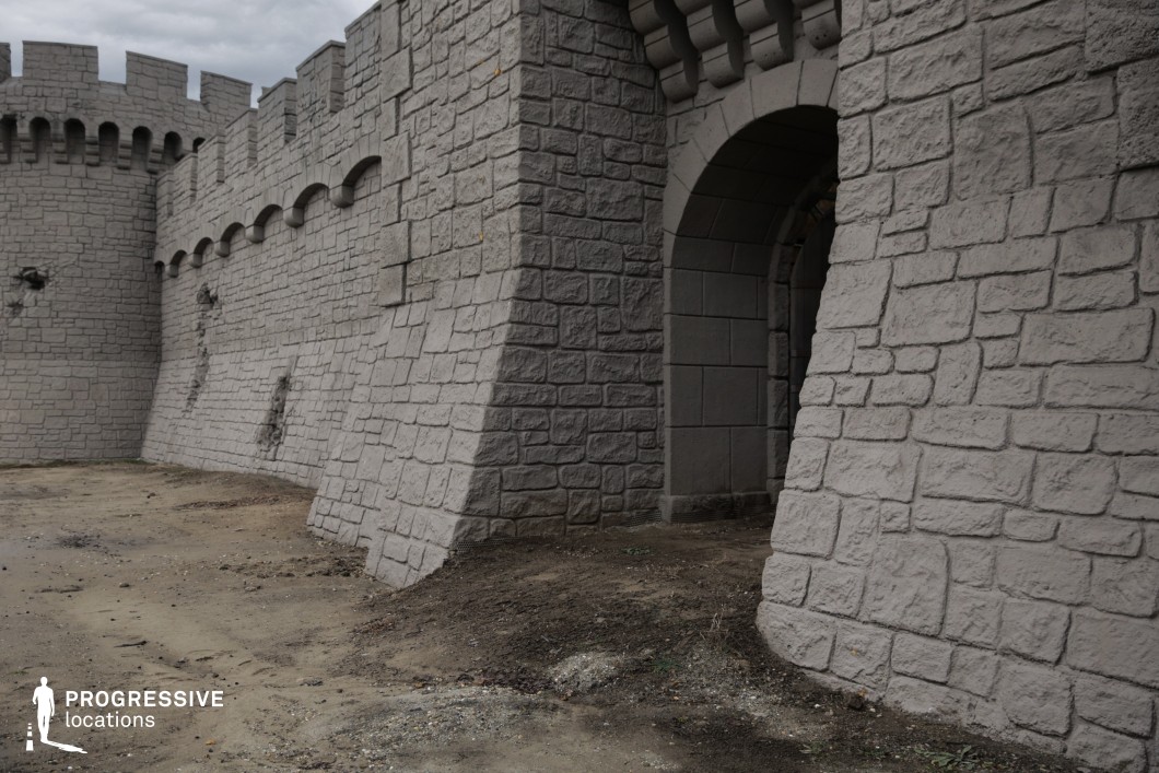 This location displays a robust stone fortification with imposing battlements, sloped base walls, and a large arched gate, evoking the outer perimeter of a medieval stronghold. The uniform grey stone texture, symmetrical crenellations, and signs of minor structural wear contribute to an atmosphere of historical defense and durability. Ideal for large-scale exterior shots, this setting is perfect for filming sieges, battle preparations, or tense negotiations at the gates in genres such as epic fantasy, medieval war films, or historical dramas.