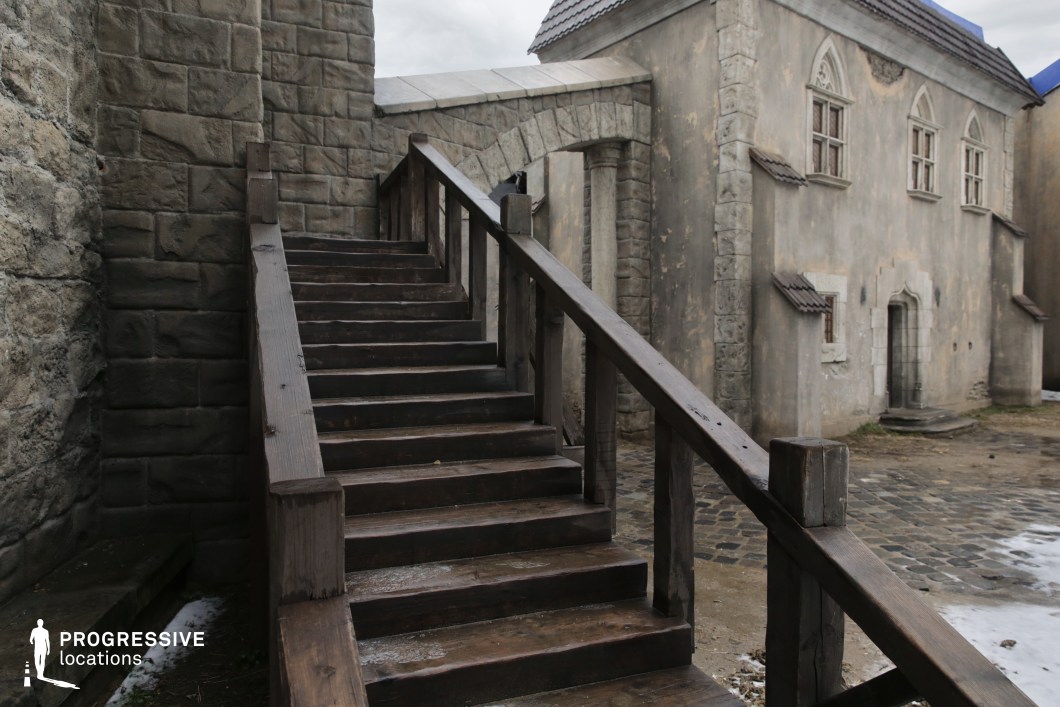 This location features a rugged medieval-style environment defined by weathered stone walls, aged plaster facades, and a robust wooden staircase leading to a narrow stone archway. The architecture combines Gothic-style window frames with fortress-like masonry, evoking a strong sense of historical depth and Old World authenticity. The scene's earthy textures and subdued tones make it ideal for period dramas, fantasy adventures, or historical action films. It serves well as a castle courtyard, town alley, or the entrance to a guild hall, offering immersive visual storytelling potential without requiring heavy set dressing.