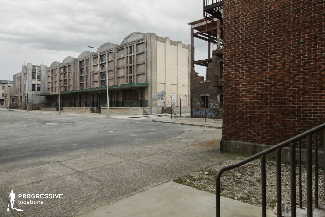 This location features a wide urban intersection dominated by weathered industrial buildings with concrete façades, repetitive vertical framing, and arched rooflines, conveying a strong sense of structural mass and industrial history. The foreground includes cracked sidewalks and minimal street elements, while the backdrop showcases exposed brickwork, chain-link fencing, graffiti-tagged surfaces, and partially collapsed or unfinished structures, enhancing the gritty, abandoned feel. The muted tones of concrete, faded paint, and oxidized metal contribute to a raw, desaturated visual palette. This versatile setting is ideal for filming dystopian, crime, or post-apocalyptic scenes, offering an atmospheric and textured environment for action or tension-driven sequences.
