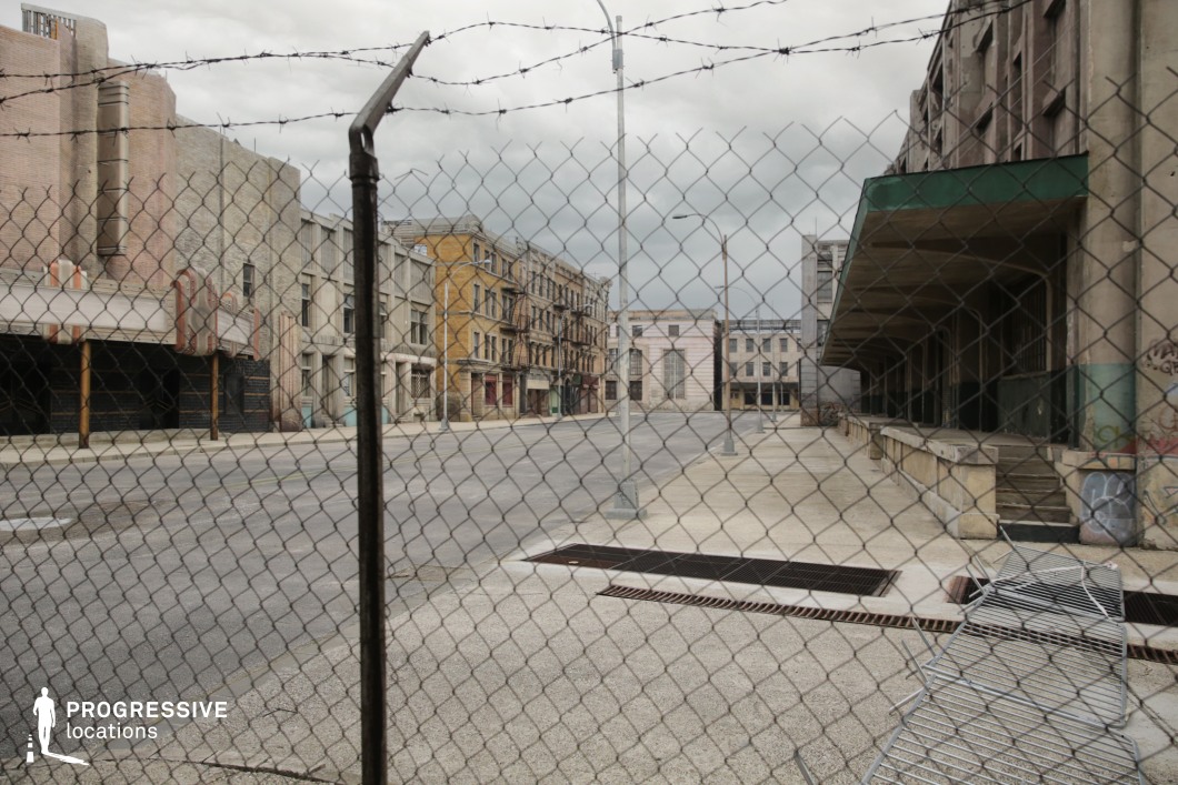 This scene presents a deserted urban street enclosed by a chain-link fence topped with barbed wire, establishing a strong sense of separation and restriction. The surrounding buildings display a mix of early 20th-century industrial and commercial architecture, including a dilapidated theater facade, apartment-like brick structures, and loading docks with concrete overhangs. The dominant materials—aged brick, concrete, and worn stucco—create a raw, textured visual environment with visible signs of decay and graffiti. The viewpoint through the fence introduces a layered composition with strong linear depth, reinforced by the symmetrical placement of streetlamps and architectural repetition. The controlled, gritty aesthetic and spatial tension make this location ideal for filming gritty crime dramas, dystopian narratives, or action sequences that require a confined, high-stakes atmosphere.