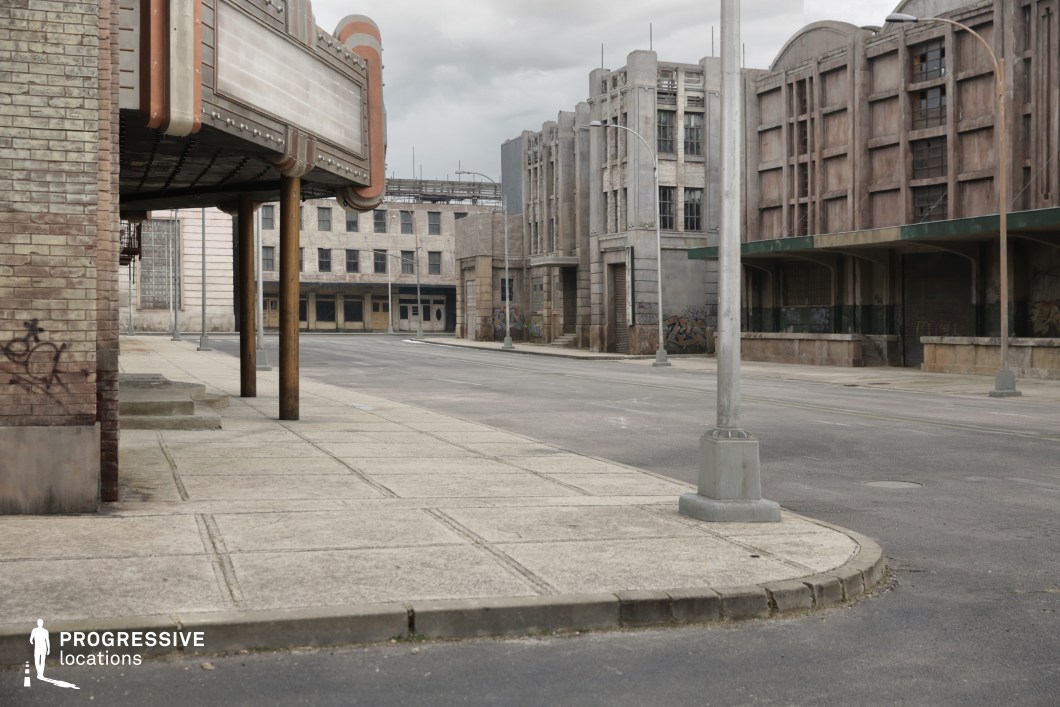 This urban streetscape features a collection of weathered, early 20th-century commercial and industrial buildings with heavy Art Deco and streamline moderne influences. The architectural style includes vertical pilasters, concrete and brick facades, and distressed detailing, enhanced by boarded-up windows and light graffiti. A vintage-style cinema marquee with retro rounded corners and marquee lighting anchors the foreground, while large-scale structures with repetitive window patterns create a rigid, symmetrical backdrop. The sidewalks are wide and defined by concrete slabs and modern streetlamps, contributing to the urban geometry. The location’s stark, desaturated tone and open layout make it especially suitable for dystopian, noir, or crime genre productions, ideal for scenes requiring desolation, tension, or stylized period atmospheres.
