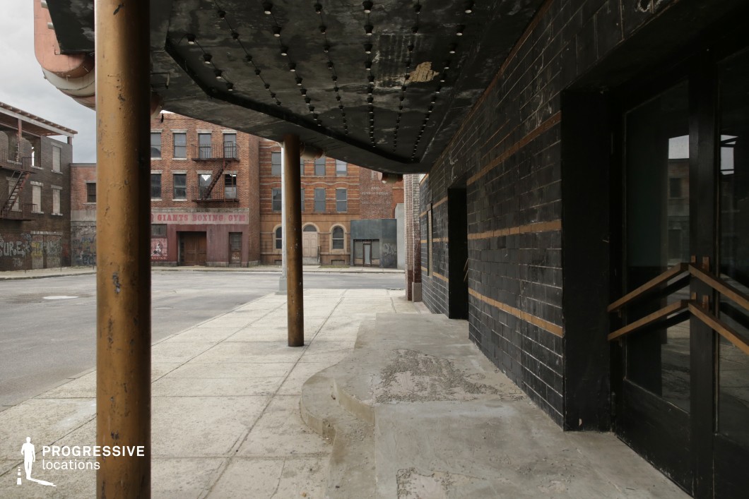 This urban street corner features a weathered building facade clad in dark, horizontally-striped tiles, with a prominent overhanging marquee supported by cylindrical metal columns. The sidewalk is wide, with uneven concrete textures and a slight curb ramp leading toward dark glass doors framed by minimalistic wooden details. Across the street, brick buildings with faded signage and fire escapes evoke a mid-20th-century industrial or downtown American atmosphere. The visual depth created by the angled perspective and the strong geometric lines enhances the gritty authenticity of the setting. This location is well-suited for film genres such as noir, crime, or urban drama, particularly for establishing exterior shots, confrontations, or transitional street scenes in a stylized city environment.