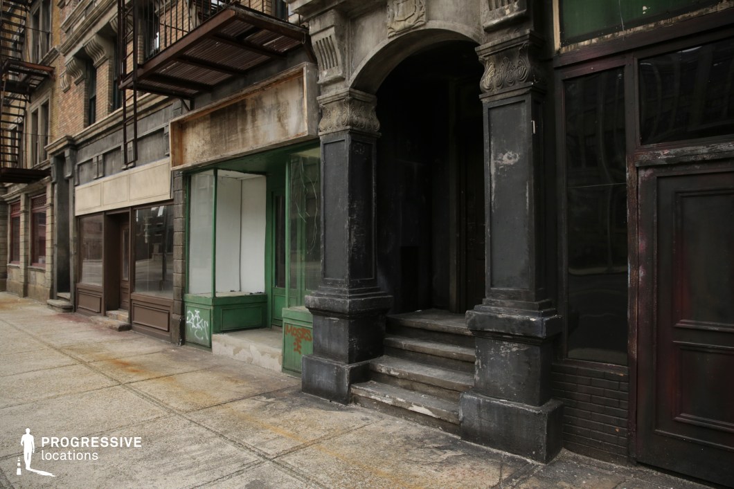 This streetscape features a row of worn, early 20th-century storefronts with a strong urban character, marked by aged textures and muted tones. Dominated by a large arched stone entrance with ornate detailing and darkened surfaces, the architecture reflects a classical revival influence juxtaposed with utilitarian commercial facades. Metal fire escapes add vertical rhythm and industrial elements above the sidewalk. The mix of faded signage areas, disused shopfronts, and graffiti-tagged panels contribute to a sense of abandonment and urban decay. This visually layered, high-contrast environment is ideal for filming crime dramas, neo-noir scenes, or character-driven narratives in dense metropolitan settings.