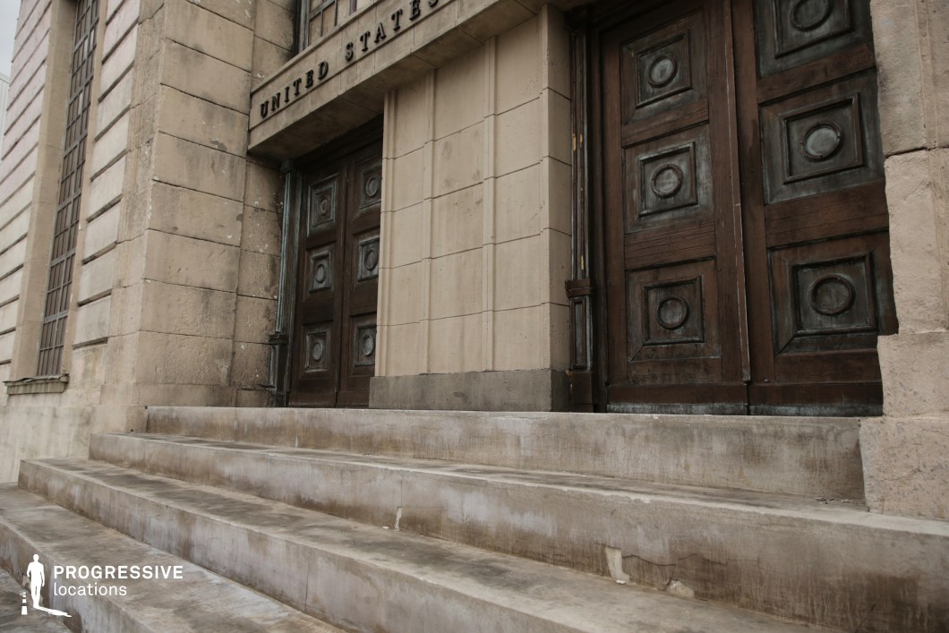 This imposing stone structure features a broad set of weathered concrete steps leading up to a pair of oversized, dark wooden doors, each embellished with deep square paneling and circular motifs. The architectural design evokes institutional authority, with thick stone blocks and vertical detailing contributing to a sense of permanence and gravity. The stone facade is punctuated by tall, narrow grid windows that allow for minimal visibility, reinforcing the austere and fortified aesthetic. The elevated angle and centered framing of the stairs emphasize symmetry and monumentality. This location is ideal for filming courtroom, political, or heist-related scenes, and fits genres such as legal drama, action thrillers, or historical fiction requiring a federal or official building exterior.