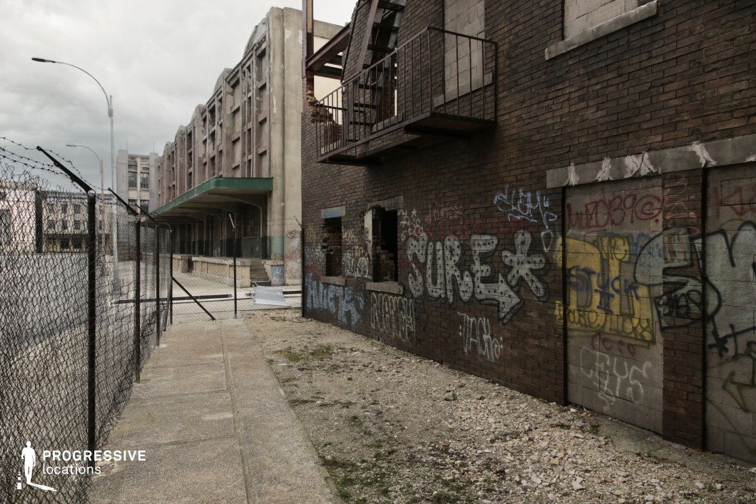 This urban alleyway is defined by a gritty, industrial aesthetic featuring aged brick walls covered in layered graffiti and faded tagging. The exposed structure includes broken windows and a rusted balcony frame, amplifying the sense of decay. Opposite, a chain-link fence with barbed wire borders the cracked concrete path, leading the eye toward a massive, monolithic warehouse structure with strong vertical lines and a green metal awning. The muted color palette and distressed materials create a raw, textured atmosphere. This setting is ideal for filming dystopian, post-apocalyptic, or crime-related scenes where an authentic sense of urban abandonment and edge is required.