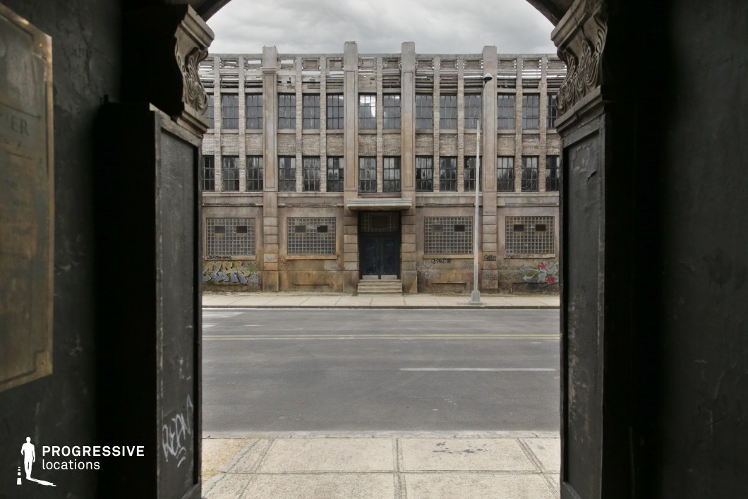 Framed by a dark archway, this view centers on a symmetrical, weathered industrial building characterized by its grid-patterned glass block windows, vertical concrete columns, and aged brickwork. The structure has a stark, fortress-like appearance, with a recessed central doorway accessed by a short flight of stairs and flanked by graffiti-marked lower walls. The facade's rhythm of vertical lines and repeated window panes contributes to its rigid, institutional aesthetic. The camera perspective through the doorway adds a layered depth, ideal for compositions involving threshold symbolism or observational framing. This location is particularly well-suited for crime dramas, dystopian thrillers, or narrative scenes requiring gritty, urban-industrial settings with architectural dominance.
