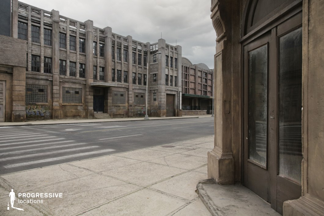 This location presents a monolithic stretch of industrial buildings with a strong Art Deco influence, marked by tall vertical lines, symmetrical window arrangements, and a repetitive concrete and brick pattern. The dominant building features thick masonry, glass block windows, and darkened entryways, evoking a sense of abandonment and functional rigidity. Foreground architectural details, such as the arched framing and worn surfaces of the nearby doorway, add depth and texture to the composition. The streetscape is wide and geometrically clean, with straight lines from the crosswalk and sidewalk amplifying the urban formality. This setting is ideal for gritty urban storytelling, including thriller, action, and post-apocalyptic genres, offering a visually rich environment for scenes requiring tension, desolation, or retro-industrial aesthetics.