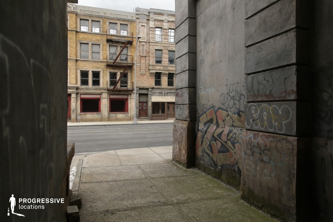 This location captures a narrow, shadowed alleyway with graffiti-covered concrete walls, leading out onto a broad urban street flanked by aged brick buildings. The alley acts as a natural frame, creating a strong contrast between the dark, enclosed foreground and the open, light-filled street beyond. The architecture across the street features early 20th-century design elements, including red-painted storefront recesses, stone detailing, and metal fire escapes that emphasize verticality and texture. The ground surfaces are worn and irregular, contributing to the raw, authentic urban character. This visually layered setting is well-suited for crime, thriller, or neo-noir genres, offering dramatic framing and a natural division of light and shadow for stylized cinematography.