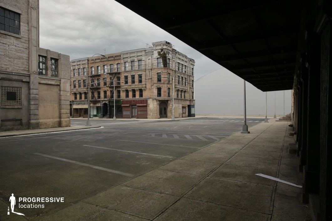 This urban location features a wide, empty intersection flanked by weathered multi-story brick buildings with boarded windows and faded facades. The architectural style suggests early 20th-century industrial or tenement structures, emphasized by visible fire escapes and ornamental stone detailing on the corner building. The cracked pavement, worn sidewalks, and minimal street furniture contribute to a stark, desolate ambiance. Strong geometric lines and the deep overhang casting shadows in the foreground add depth and contrast to the visual composition. This setting is particularly suited for filming gritty dramas, noir-inspired thrillers, or dystopian urban scenes, offering a controlled, atmospheric environment with strong architectural character.