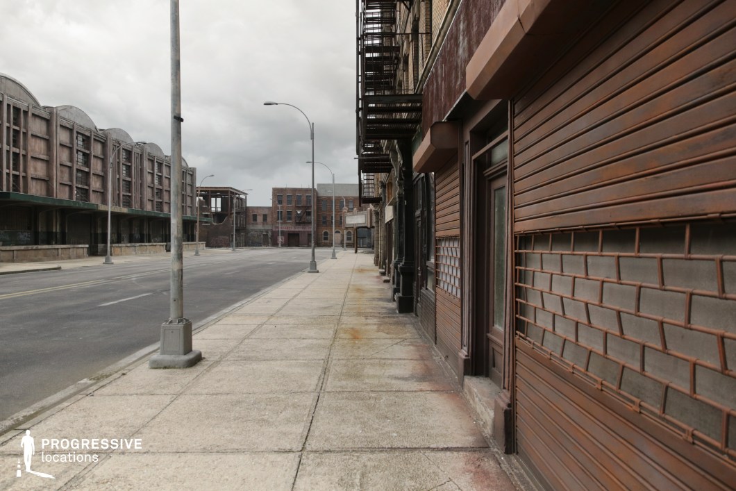 This location showcases a wide, linear urban streetscape defined by industrial and early 20th-century architectural elements. The right side features aged brick façades with metal roll-up shutters, fire escapes, and dark storefronts that suggest former commercial or service use. On the left, a long row of concrete and brick warehouses with large arched roofs and covered loading docks adds a sense of scale and historical utility. The entire setting is built around hard, weathered materials—such as steel, concrete, and aged brick—producing a stark, utilitarian aesthetic. The uninterrupted sidewalks, symmetrical lamp posts, and distant brick façades contribute to a cinematic depth. This location is ideal for crime dramas, dystopian thrillers, or period pieces requiring an industrial or economically challenged urban environment.