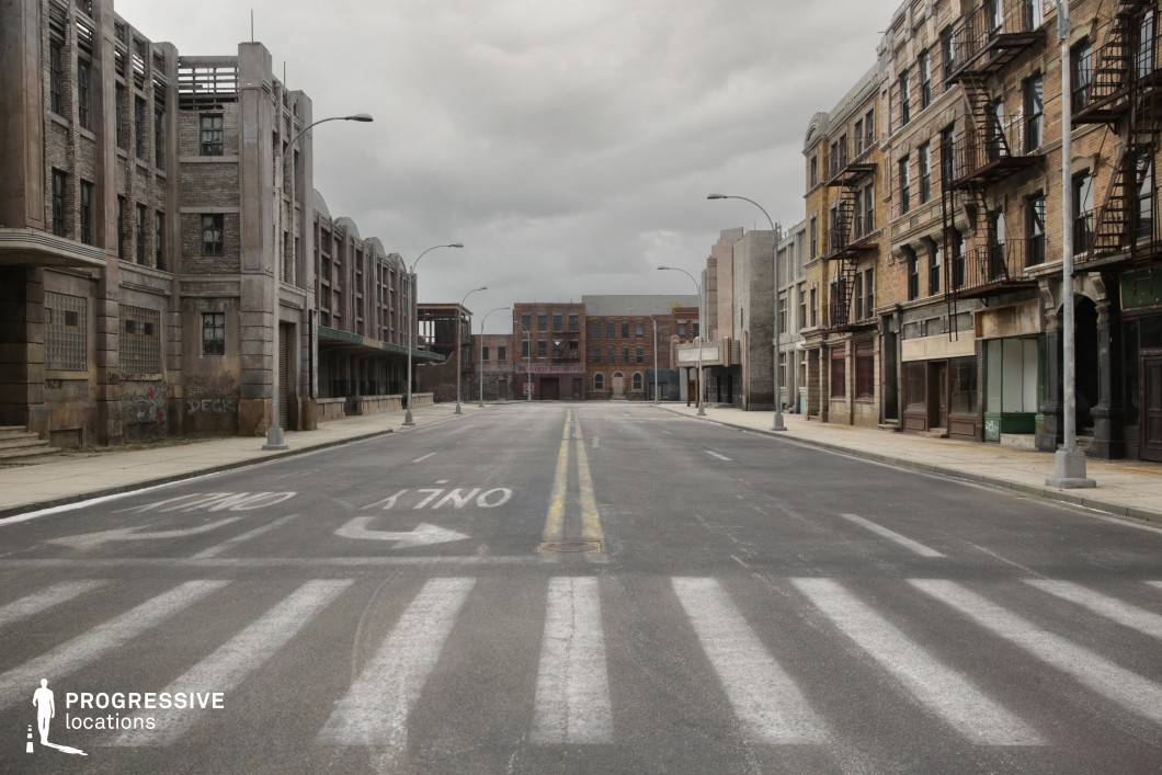This wide street scene features symmetrical rows of early 20th-century style buildings with worn brick façades, repetitive window arrangements, and external fire escapes. The architecture on both sides blends industrial and residential characteristics, marked by tall ground-floor shopfronts and multi-story apartment structures above. The asphalt road, complete with directional street markings and aligned streetlights, emphasizes the linear depth and vanishing point, enhancing the scene's sense of scale. With its aged textures, faded signage, and theatrical emptiness, the location is highly versatile for filming urban decay, crime dramas, or retro city environments. It is especially suitable for action sequences, chase scenes, or establishing shots in thrillers and noir-style narratives.