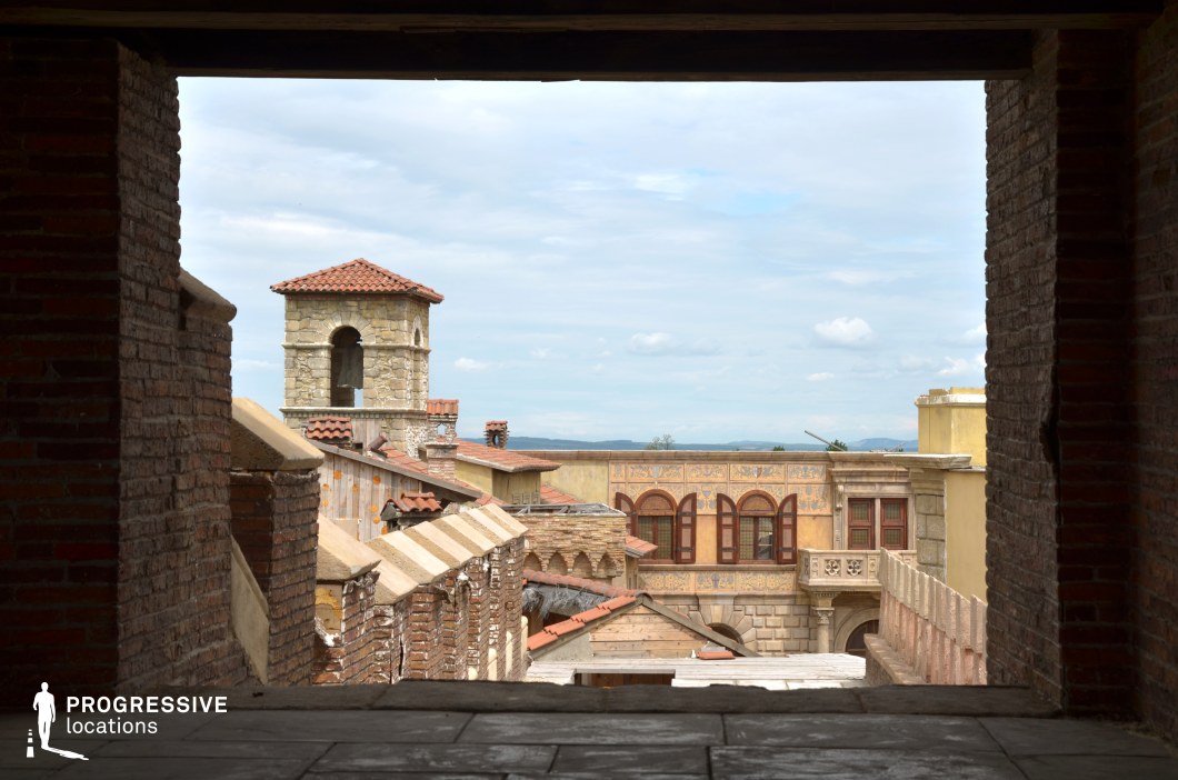 This elevated perspective captures a layered architectural ensemble characterized by stone and brick structures with Mediterranean influences. Dominated by a central bell tower with a red-tiled roof and rough-hewn stonework, the scene is framed by decorative façades featuring ornate cornices, arched windows with wooden shutters, and pastel-colored plaster. Foreground brickwork and stair-like elements lead the viewer’s eye toward the more distant buildings, establishing a rich sense of depth. The harmonious mix of textures—stone, brick, and plaster—combined with traditional tile roofing creates a cohesive, timeless aesthetic. The framing through a shadowed archway adds a cinematic vignette to the composition. This location is especially suitable for historical dramas, Renaissance-era narratives, or fantasy films requiring an authentic Mediterranean-style townscape with visual layering and architectural character.
