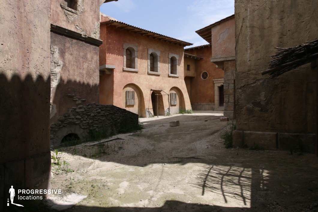 This compact alleyway reveals an intimate view of sun-drenched, earth-toned façades with textured plaster walls, stone accents, and wooden shutters. The architectural simplicity is enhanced by rounded archways, modest detailing, and irregular surface treatments, evoking a timeless rural or small-town Mediterranean environment. The interplay of shadow and light creates depth and atmosphere, with natural textures contributing to a sense of place. This location is ideal for filming historical dramas, coming-of-age stories, or rustic adventure tales. It serves well for quiet character interactions, secretive meetings, or moments of reflection within a convincingly historic Southern European setting.