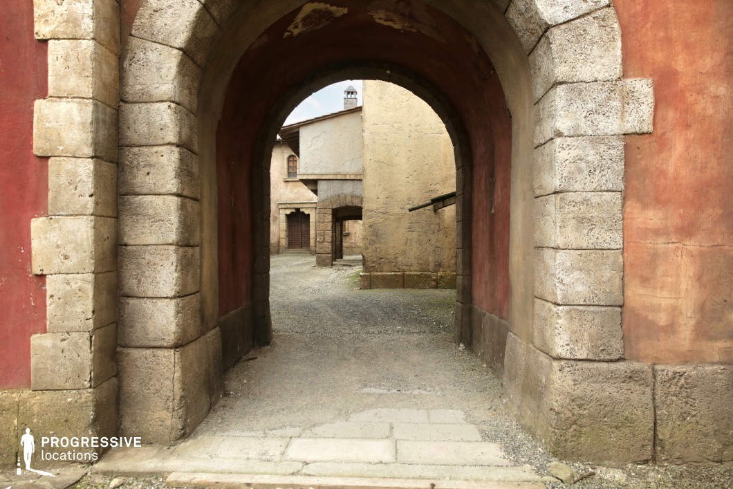 This narrow alleyway is framed by a rounded stone archway leading into a compact courtyard of rustic, textured buildings. The architecture is characterized by coarse stucco facades in muted ochre and terracotta tones, with exposed stone block details at the base and corners. The layered perspective reveals multiple architectural arches and recesses, creating strong depth and geometric repetition. Weathered surfaces and uneven gravel ground add an authentic, aged quality that suggests historical or rural European architecture. This intimate, enclosed setting is ideal for scenes requiring a sense of seclusion, tradition, or timelessness—well-suited for period dramas, historical reenactments, or character-driven narratives set in old-world environments.