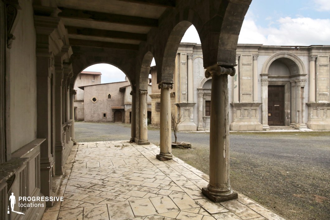 This architectural location features a stone colonnade with rounded Romanesque arches and classical columns forming a sheltered walkway that opens into a spacious courtyard. The foreground showcases patterned stone flooring, while the backdrop consists of a blend of Renaissance-style and rustic buildings with plastered façades, arched doorways, and decorative cornices. The symmetry of the arcades and the repetition of classical elements like pilasters and columns establish a refined historical aesthetic. The space is defined and enclosed by surrounding structures of varying textures and elevations, offering visual depth and architectural contrast. The balanced composition and timeless design make this an excellent setting for historical dramas, period pieces, or scenes requiring a European old-town atmosphere with cinematic architectural framing.