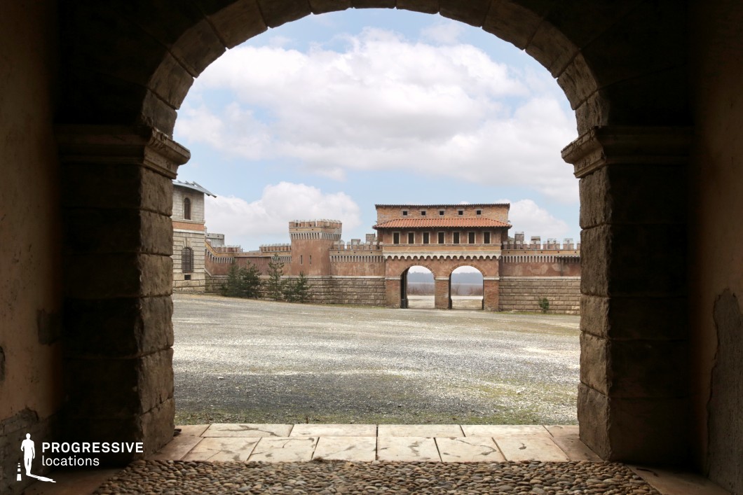 This expansive fortress courtyard is framed by a stone archway in the foreground, offering a dramatic visual gateway to the imposing gatehouse beyond. The gatehouse features symmetrical twin arches and a crenellated parapet, with a terracotta-tiled roof and small, regularly spaced windows that emphasize its defensive yet stylized character. The enclosing walls and towers, constructed from weathered stone and brick, reinforce a medieval or Renaissance architectural theme, while the spacious gravel courtyard allows for dynamic movement and staging. The clear sightlines and layered depth created by arches and perspective make this location especially cinematic. It is well-suited for historical epics, fantasy productions, or period war dramas, offering a grand and immersive environment for large-scale exterior scenes.