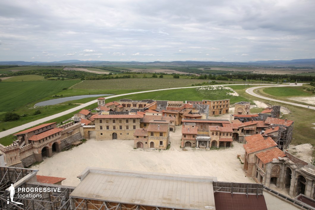 Captured from an elevated vantage point, this image reveals a large-scale historical backlot designed to resemble a Mediterranean or Southern European fortified town. The arrangement includes sandstone-colored buildings with terracotta tile roofs, colonnades, archways, and plazas, forming a coherent and immersive environment. The backdrop features expansive open fields, vineyards, and gentle hills, creating a striking contrast between the architectural density and natural openness. Scaffolding and set structures along the periphery indicate its functional purpose as a cinematic environment. The symmetrical layout, spacious central courtyard, and surrounding rural landscape make this location exceptionally well-suited for epic period productions, historical reconstructions, or scenes requiring large-scale outdoor environments with architectural authenticity.