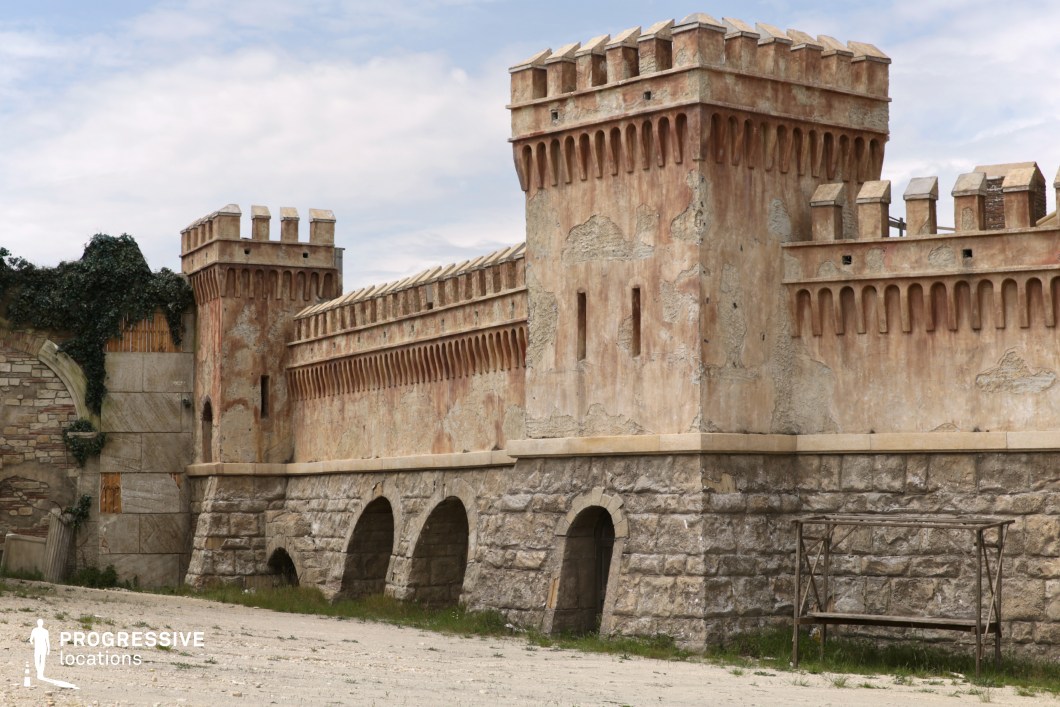 This imposing medieval-style location features a long, elevated stone wall flanked by tall square towers, constructed with a combination of rough-hewn stone at the base and aged, textured plaster on the upper sections. The design includes battlements, slit windows for archers, and evenly spaced corbels beneath the parapets, reinforcing its fortified appearance. The arched passageways at ground level suggest a defensive structure with potential functional depth, while the subtle signs of wear and discoloration add to its historical character. The composition is symmetrical and horizontal, captured from a frontal ground-level perspective that emphasizes the linear continuity of the wall. This site is ideal for fantasy, historical, or epic genre productions, offering a visually strong, enclosed backdrop for battle preparations, fortress exteriors, or period transitions.