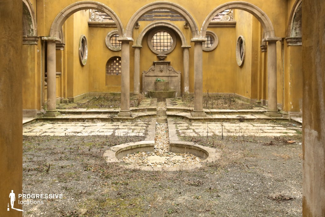 This enclosed courtyard features a symmetrical layout framed by a series of classical stone arches supported by cylindrical columns, evoking a Renaissance or Mediterranean architectural influence. The ochre-toned walls, round windows, and recessed geometric detailing create a warm and aged ambiance. At the center stands a dry, decorative fountain with a carved basin and emblematic relief, surrounded by a grid of overgrown stone paving and gravel pathways. The structure’s spatial symmetry and aged surfaces offer a textured visual environment, with natural decay contributing to its atmospheric charm. This location is ideal for period pieces, contemplative dramas, or fantasy productions seeking an intimate, historically inspired setting with visual depth and architectural character.