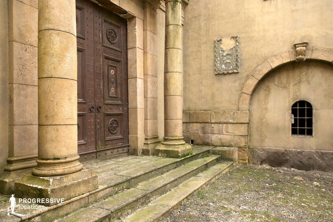 This setting presents a grand stone entrance framed by two substantial cylindrical columns and a series of worn, moss-lined steps, conveying a historic and formal atmosphere. The heavy, dark wooden double doors feature intricate relief work and metal studs, reinforcing the sense of permanence and age. The adjacent wall includes a barred arched window and a partially damaged stone crest, contributing to the weathered, institutional character of the space. The surfaces are primarily composed of sandstone and aged plaster with subtle discoloration and surface erosion, enhancing the visual texture. The overall layout is symmetrical and architecturally balanced, with a neutral, monochromatic palette dominated by beige and brown tones. This location is highly suitable for historical, religious, or dramatic productions, offering an authentic backdrop for scenes requiring solemnity, tradition, or architectural gravitas.