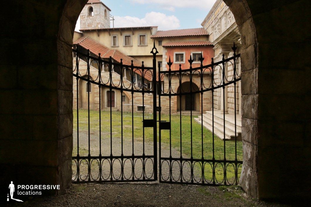 This setting captures a symmetrical view through a large stone archway, framed by an ornate wrought iron gate that opens onto a courtyard enclosed by Mediterranean-style buildings. The architecture combines stone and stucco facades with terracotta tile roofing and arched colonnades, evoking a Southern European aesthetic. The courtyard surface alternates between gravel and patchy grass, adding a natural, slightly aged texture. Architectural elements such as narrow windows, stone stairs, and varied elevations provide visual depth and historic character. This location is well-suited for historical, dramatic, or ecclesiastical scenes, offering a secluded, cinematic courtyard perfect for controlled, focused sequences in film and television.