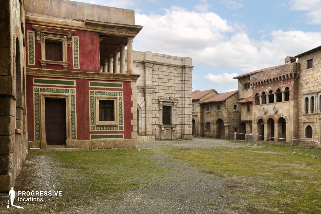 This open square is enclosed by a variety of stone and plaster buildings reflecting a blend of Romanesque and classical influences. On the left, a striking red façade with green ornamental patterns and stone-framed windows contrasts with the surrounding muted stone structures. To its right, a monumental grey stone building with pilasters and detailed masonry adds a formal, institutional tone. Further along, a series of arched arcades and brick decorations introduce a rhythmic, medieval aesthetic. The ground is a mix of grass and gravel, enhancing the historical authenticity and offering versatility for production design. This visually rich and stylistically varied location is well-suited for epic dramas, historical narratives, or fantasy genres that require layered architectural depth and a convincingly aged urban square.