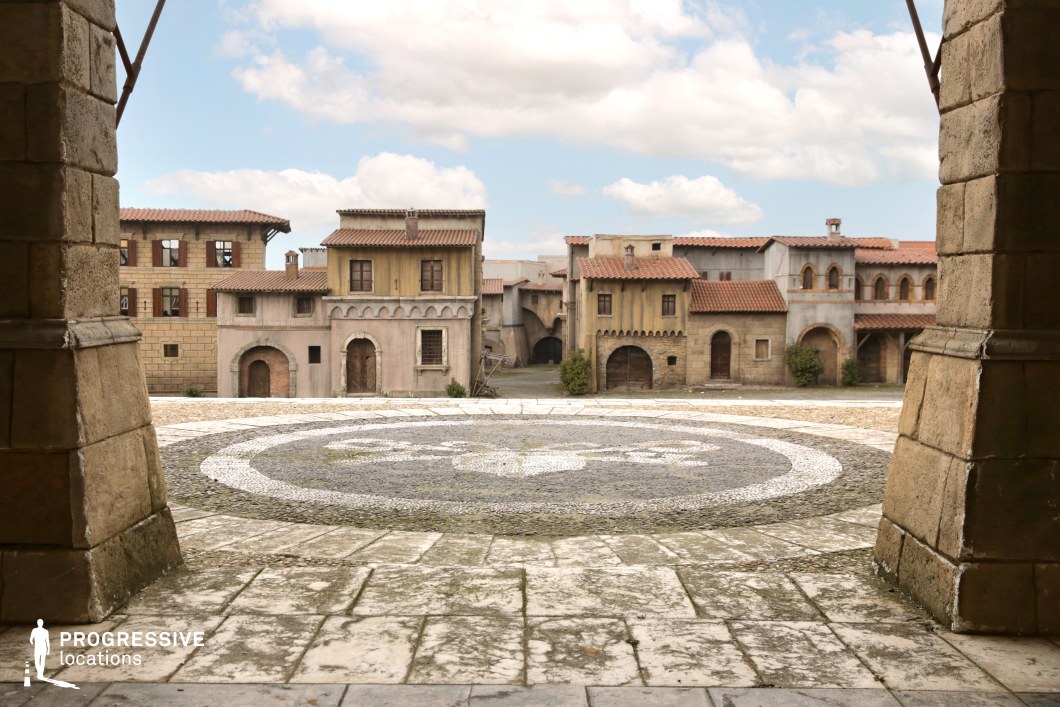 This location presents a stylized Renaissance square with a central circular mosaic pattern set into a wide cobblestone surface, surrounded by rustic façades featuring stucco finishes, arched entrances, and terracotta roof tiles. The architecture reflects a blend of Italianate and Spanish colonial influence, characterized by small balconies, arcades, and vertically compact window arrangements. The buildings vary in color from pale beige to warm ochre and weathered grey, creating a textured, cinematic backdrop with historical undertones. The framing foreground columns enhance depth and symmetry, making the open square visually balanced and compositionally strong. As a purpose-built film set, it offers controlled conditions and an immersive, old-world atmosphere ideal for shooting historical epics, period adventures, or fantasy productions requiring a European village environment.