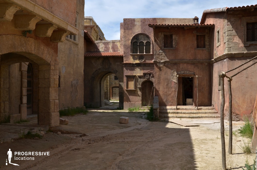 This narrow set recreates an old Mediterranean-style street, enclosed by irregular plaster façades in earthy tones of terracotta and sandstone. The surfaces show deliberate signs of weathering and wear, enhancing the realism of the historical aesthetic. Key architectural features include arched doorways, small wooden shutters, decorative grilles, and red-tiled roof overhangs. A vaulted stone archway at the end of the alley creates depth and visual layering, while uneven ground textures and stone steps add dimensionality to the space. The asymmetrical arrangement of structures and limited open sky frame a convincingly aged, intimate environment. Ideal for historical, biblical, or fantasy productions, this location supports immersive storytelling that demands a convincingly ancient urban setting with strong textural and spatial character.