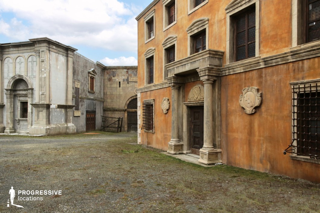 This film set captures the essence of a Mediterranean or Southern European urban corner, defined by a dominant terracotta-toned building with classical stone detailing. The façade includes pilasters, ornamental reliefs, and window moldings that suggest a historical civic or noble structure. Opposite, a weathered pale grey structure features arched elements and textured stonework, contributing to an aged, authentic ambiance. The worn plaster, decorative coats of arms, and iron-barred windows reinforce the visual narrative of historical depth. The open, gravel-covered space between the buildings offers an unobstructed staging area with controlled sightlines and versatile movement potential. This set is particularly effective for historical dramas, Renaissance-inspired scenes, or fantasy productions seeking a convincingly aged urban setting with architectural grandeur.