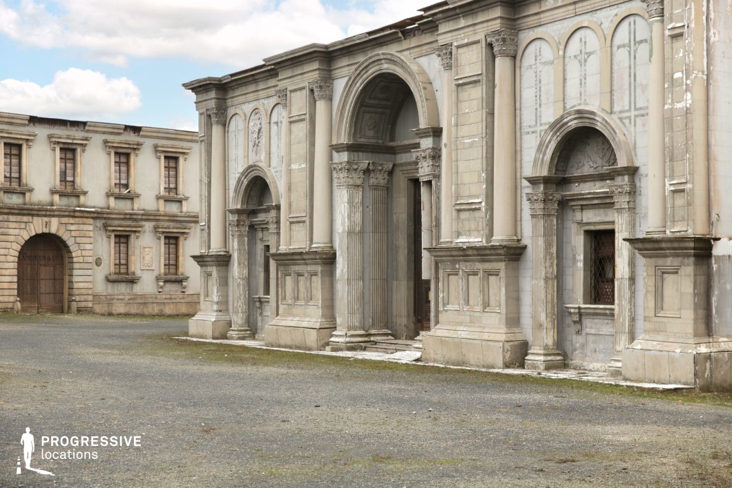 This location presents a pair of grand neoclassical-style facades, dominated by tall Corinthian columns, rounded and rectangular archways, and decorative relief panels. The stone surfaces exhibit weathered textures and slight wear, adding a sense of age and realism. The architectural rhythm is enhanced by repeated window frames and pilasters, giving the structure monumental symmetry. The open gravel square in front offers unobstructed space for camera movement and staging. This environment is especially suitable for historical, political, or epic genres, serving well as a courthouse, palace entrance, or other formal institutional setting due to its imposing scale and classical detailing.