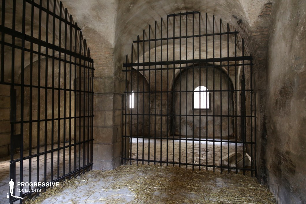 This enclosed space resembles a historical prison cell, featuring thick stone walls, arched ceilings, and heavy vertical iron bars dividing the chambers. The architecture includes a series of rounded vaults and deep-set windows that permit limited natural light, casting shadows across the rough stone and plaster surfaces. The flooring is composed of worn stone tiles scattered with straw, contributing to a raw, austere ambiance. The ironwork is dominant and rhythmically structured, enhancing the feeling of containment and tension. The combination of robust masonry, aged surfaces, and minimal lighting creates a visually impactful, claustrophobic atmosphere. This location is ideal for filming historical dramas, medieval or fantasy productions, or any genre requiring authentic dungeon, captivity, or fortress interior scenes.