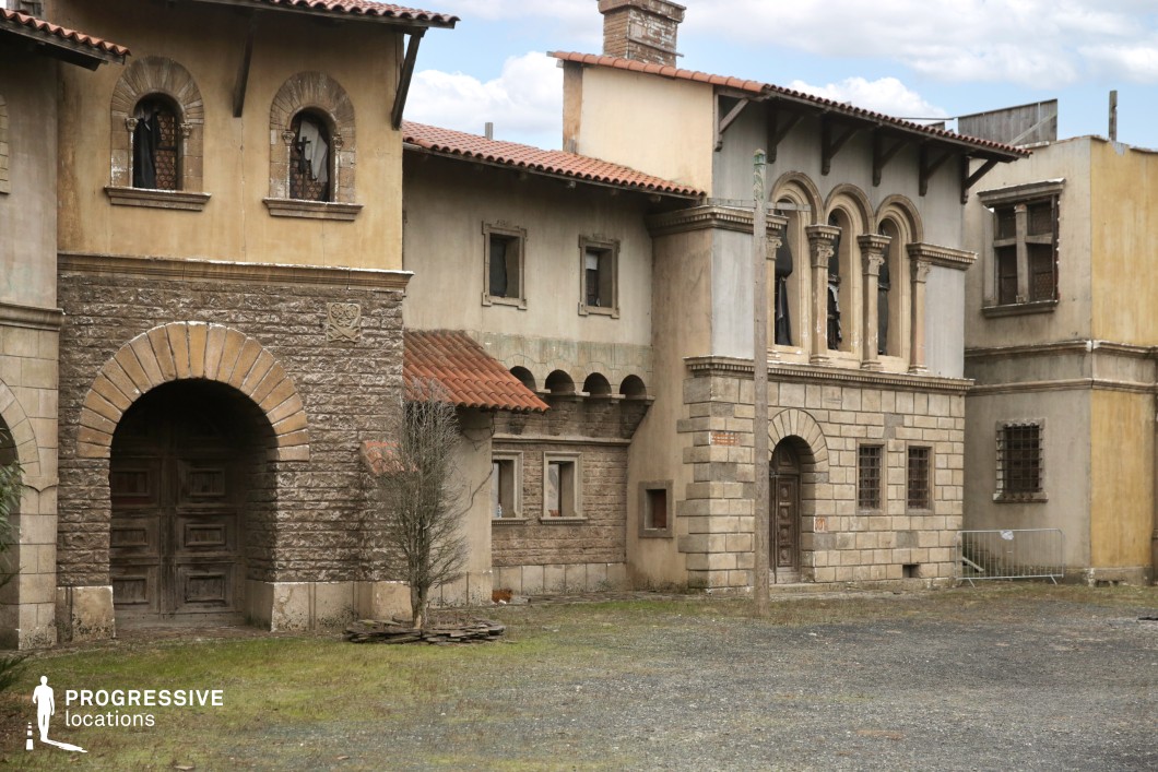 This scene captures a compact grouping of Mediterranean-style buildings characterized by stone archways, red-tiled roofs, and decorative elements such as columned windows and rustic textures. The facades show intentional signs of wear, enhancing the authenticity and historical depth of the setting. The mix of architectural details—from carved lintels to patterned stone bases—suggests a once-prosperous neighborhood now faded with time. This location is ideal for filming historical dramas, mafia stories, romantic tragedies, or fantasy sequences. It provides an evocative backdrop for scenes involving intrigue, neighborhood tension, or intimate storytelling in a timeless southern European atmosphere.