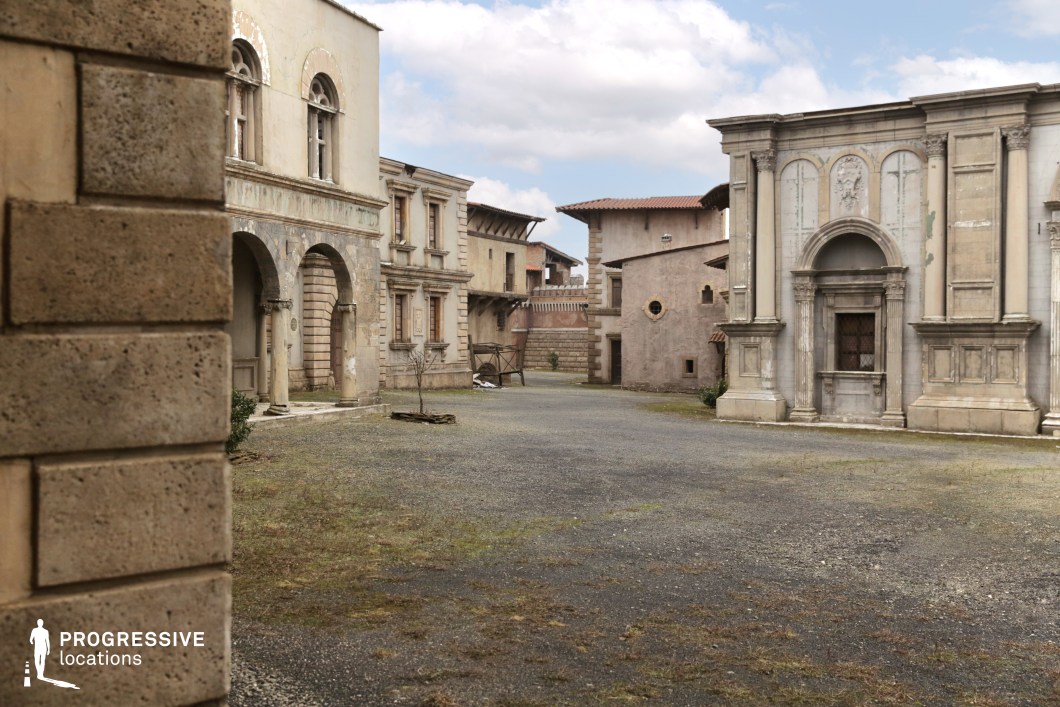 This location presents a quiet, enclosed courtyard framed by historical-style buildings with varied classical architectural details, including arched colonnades, pilasters, and stuccoed facades in muted beige and grey tones. The structures exhibit visible aging such as surface wear, flaking plaster, and faded stonework, adding a sense of authenticity and temporal depth. The buildings vary in design, combining elements of Renaissance and rustic Mediterranean styles, with high-set windows, ornamental reliefs, and terracotta roofing. The central open space is surfaced with a mix of gravel and sparse vegetation, enhancing the unrefined, timeworn character. The narrow street openings and surrounding architecture form a visually contained environment, making the location well-suited for period pieces, historical dramas, or European-themed productions. Its balanced mix of elegance and decay allows for flexible adaptation in both narrative and stylistic filming contexts.