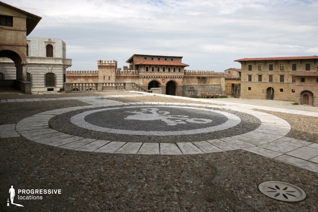 This expansive open-air location features a fortified square surrounded by renaissance-influenced stone structures with symmetrical facades, arched entrances, and terracotta roof tiles. The ground is covered in cobblestones and white stone tiling, with a large circular mosaic at the center that creates a strong visual focal point. The perimeter walls include battlements and decorative stone railings, while the backdrop showcases a fortified gatehouse flanked by towers and a series of upper-level windows. The uniformity in materials—stone, plaster, and terracotta—creates a cohesive historical atmosphere, while the wide, open layout allows for dynamic camera movement and clear spatial orientation. The location is ideal for filming historical dramas, fantasy epics, or period adventures, offering a controlled yet authentic-looking environment reminiscent of Mediterranean or early European cities.