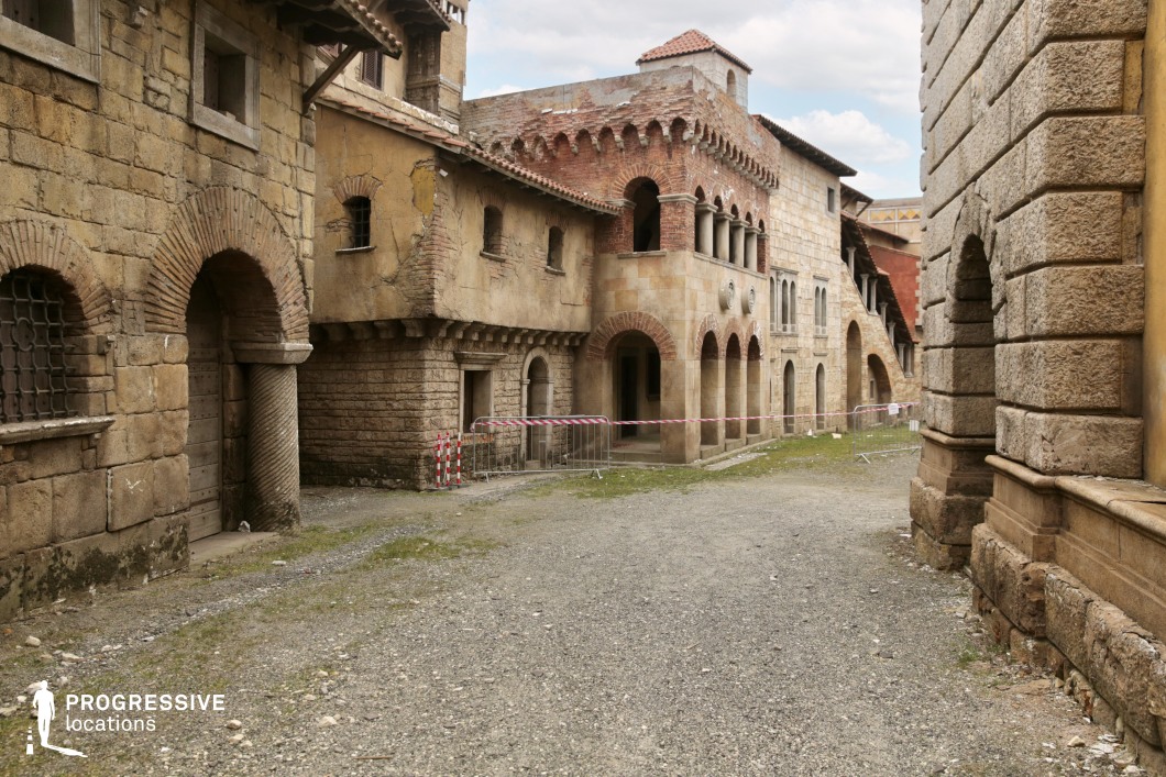 This enclosed alleyway features robust stone and brick buildings with a distinctly medieval character, defined by arched doorways, columned arcades, and deeply set windows. The façades display a mix of coarse-hewn blocks, red brickwork, and aged plaster, creating strong visual texture and historic ambiance. Architectural details like corbels, decorative friezes, and barred windows contribute to an impression of fortified structures, while the uneven gravel street adds a raw, grounded quality. The narrow spatial proportions and earthy color palette reinforce a sense of enclosed antiquity. This location is particularly well-suited for historical or fantasy genres, offering a dramatic and immersive setting for scenes requiring a fortified urban backdrop or medieval ambiance.