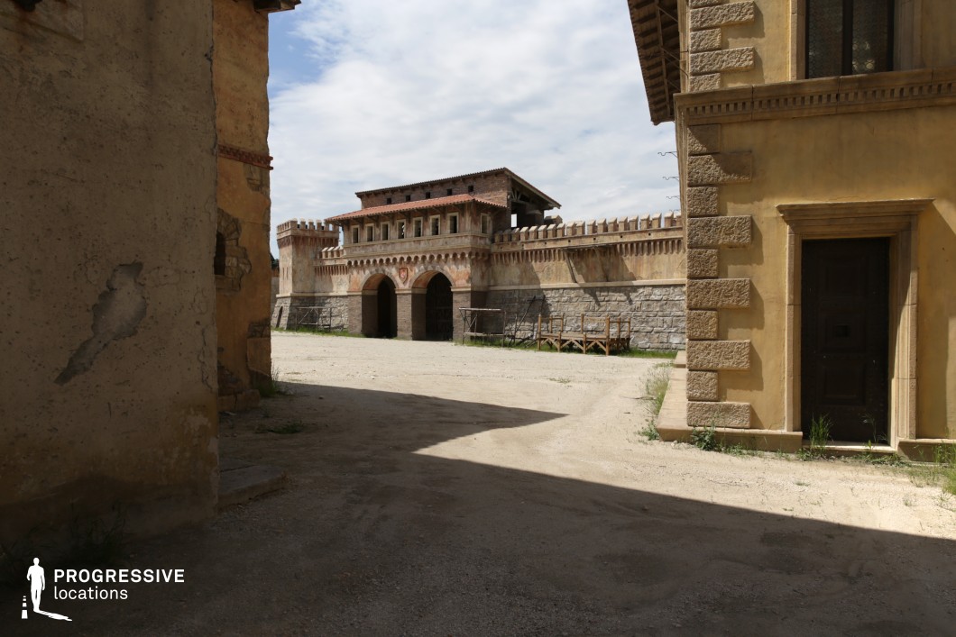 This location showcases a fortified structure with medieval-inspired architecture, framed by textured, ochre-colored buildings in the foreground. The dominant visual element is a castle-like gatehouse featuring a dual-arched entrance, battlements, and a rhythmically spaced window line, all set atop a stone base. The surface of the courtyard is covered with pale, compacted gravel, complementing the earth-toned masonry. Architectural elements such as quoined corners, wooden doorways, and visible roof beams enhance the historic atmosphere. The scene is defined by strong geometric lines and the interplay of sunlit open space with shaded passageways. Ideal for filming historical, fantasy, or period adventure genres, this setting offers a versatile backdrop for scenes requiring fortified towns, citadels, or old-world streetscapes.
