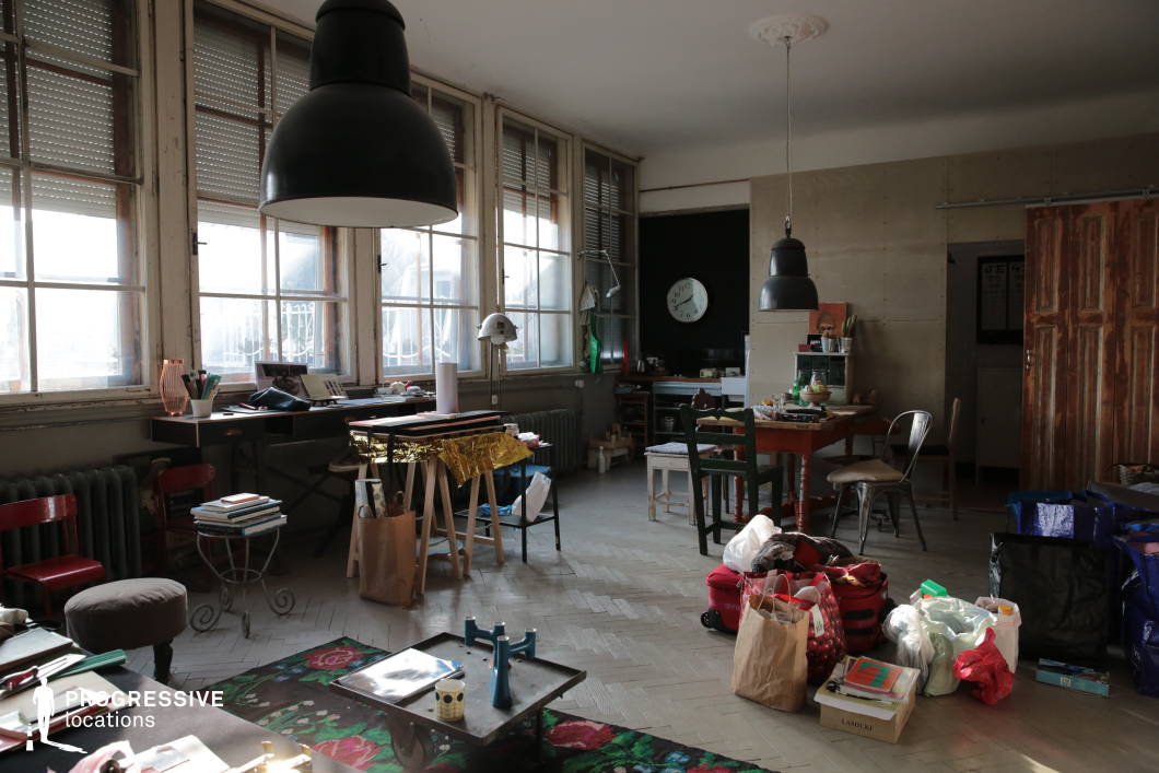 This spacious artist's studio combines industrial and vintage aesthetics, characterized by large multi-pane windows with aged wooden frames, which allow diffused natural light to filter in. The high ceiling is punctuated by oversized black pendant lamps, reinforcing the industrial ambiance. The room features an eclectic mix of furniture, including mismatched chairs, rustic wooden tables, and metal stools, all contributing to a creative and lived-in atmosphere. The floor is composed of light herringbone tiles, adding texture and contrast to the raw, unfinished walls. A workspace cluttered with art supplies, books, and personal objects creates an authentic and dynamic environment, ideal for capturing a bohemian or artistic lifestyle. Various bags and storage containers suggest a transient or evolving creative process, making the space suitable for documentary-style shoots, character-driven storytelling, or editorial photography.